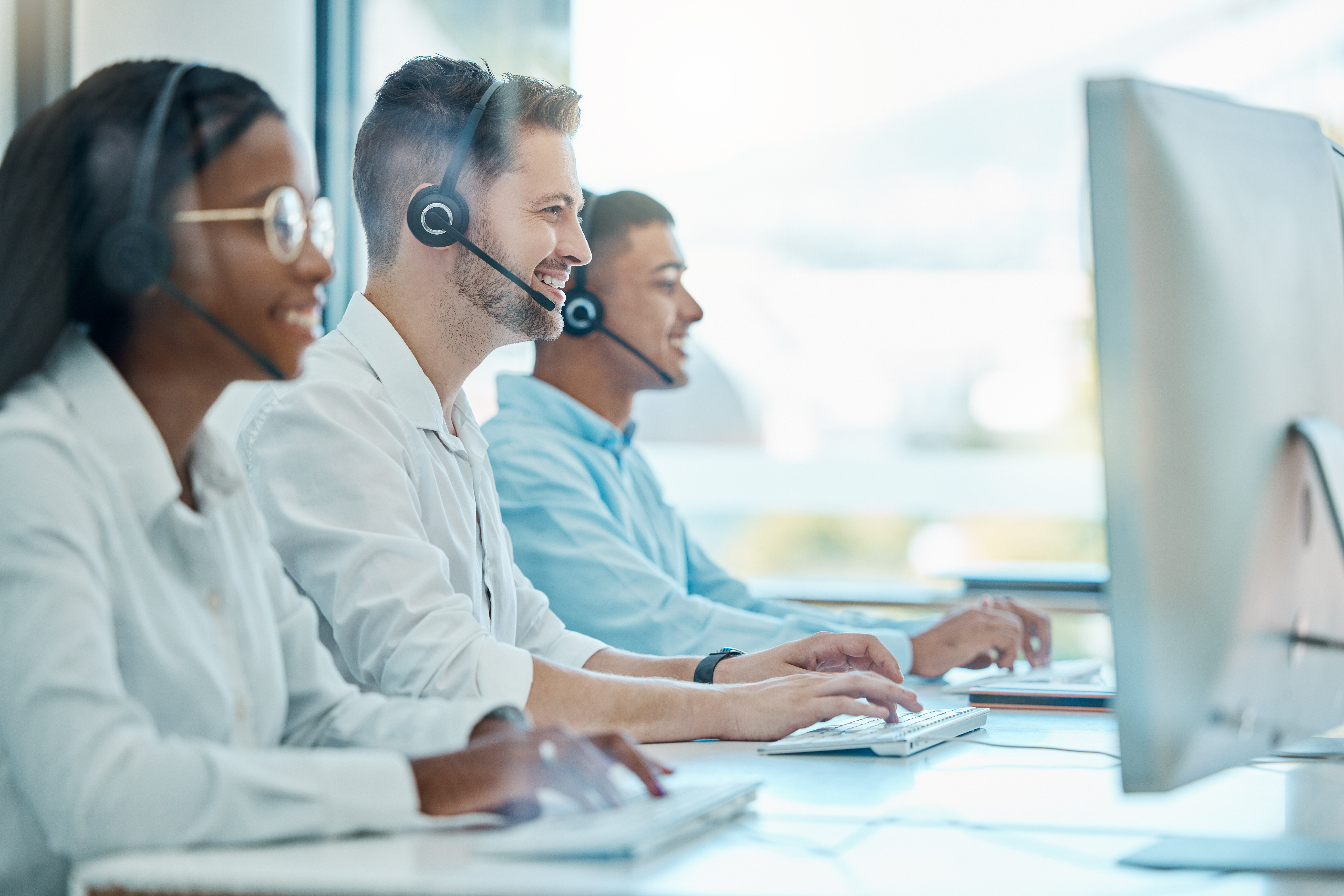 Three customer service representatives wearing headsets are smiling and actively engaged at their computers in a bright, modern office setting. The male representative in the foreground and the two diverse colleagues in the background illustrate a dynamic and professional Business Development Center (BDC) at an automotive dealership, focusing on customer interaction and support.