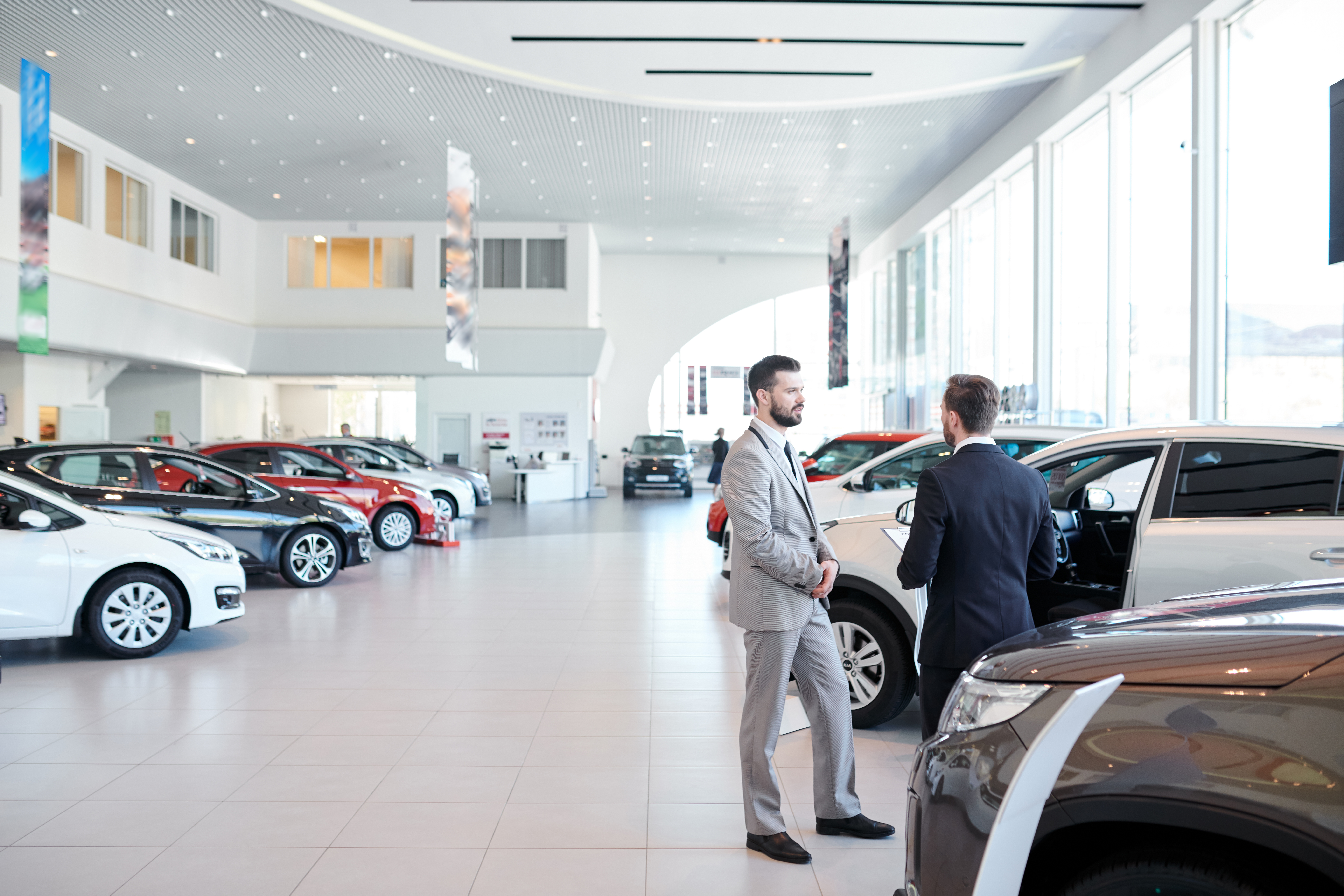 Two sharply dressed sales professionals discussing performance on a bright car dealership floor, reminiscent of dealerships that have improved performance and customer satisfaction with employee scorecards. Their earnest conversation mirrors the collaborative approach that contributes to a dealership's success and positive customer experiences.